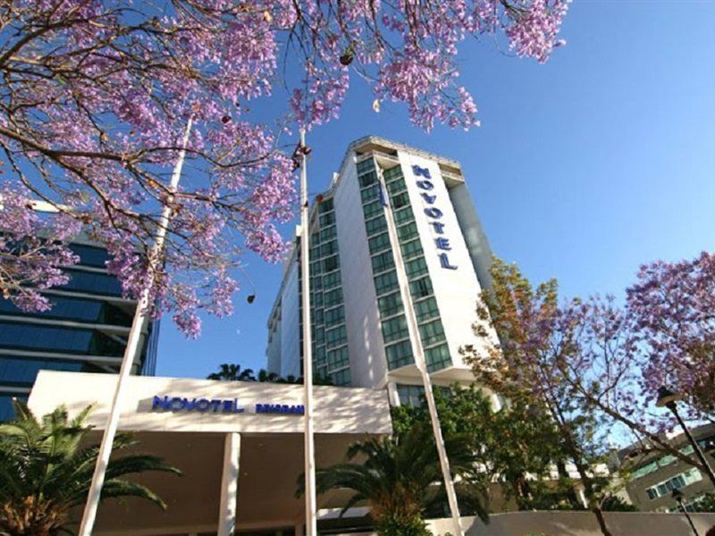 อโมรา บริสเบน Hotel ภายนอก รูปภาพ The photo shows a modern hotel building, identified as "Novotel," rising above a landscaped area with palm trees. In the foreground, there are vibrant purple flowering trees, likely jacarandas, which add to the picturesque scene. The sky is clear and